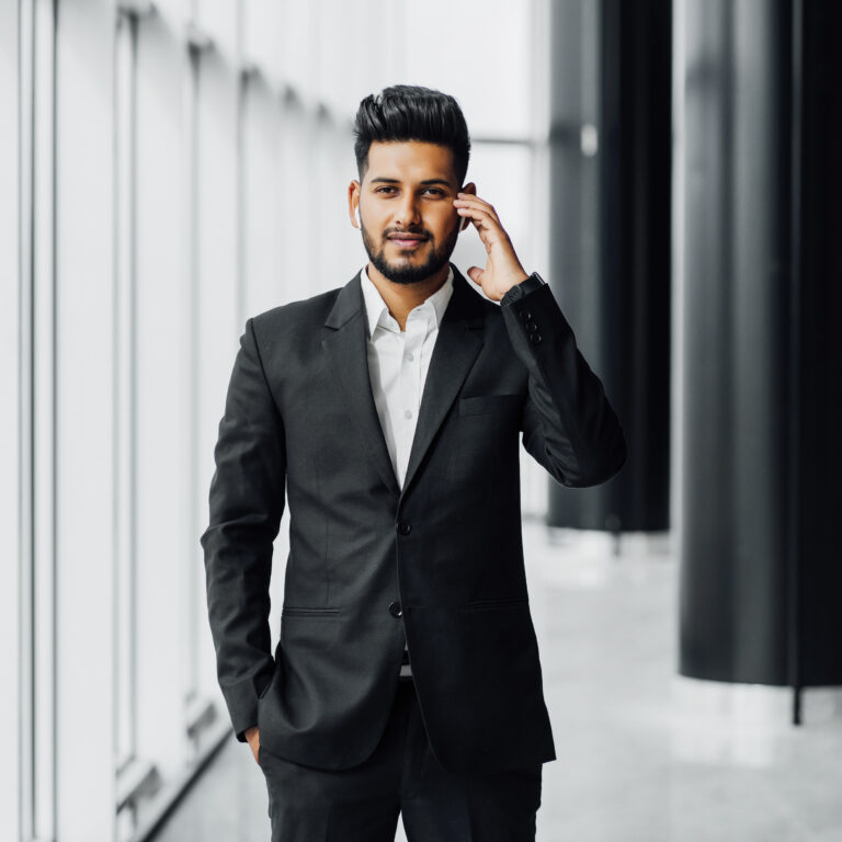 An attractive bearded Indian businessman in a modern office center, wearing a black suit, holds his hand near a wireless earphone and looks at the camera. Young Indian businessman.